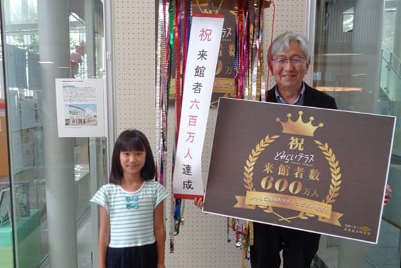 600万人達成のくす玉を後ろにした達成者の子どもとと図書館長の記念写真
